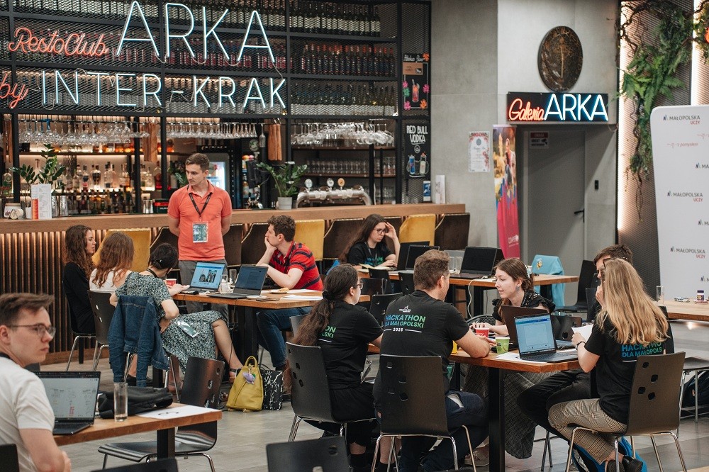 Widok sali na kt&oacute;rej studenci pracują nad wyzwaniami. Z tyłu sali stoi mentor i pomaga jednemu z zespoł&oacute;w.