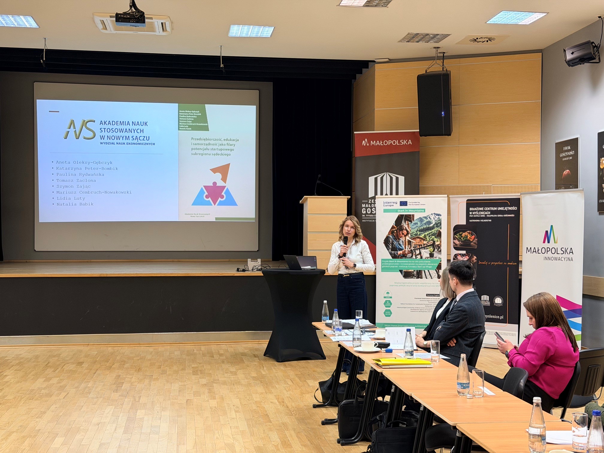 Kobieta w białej bluzce przedstawiająca prezentację na rzutniku. W tle roolupy