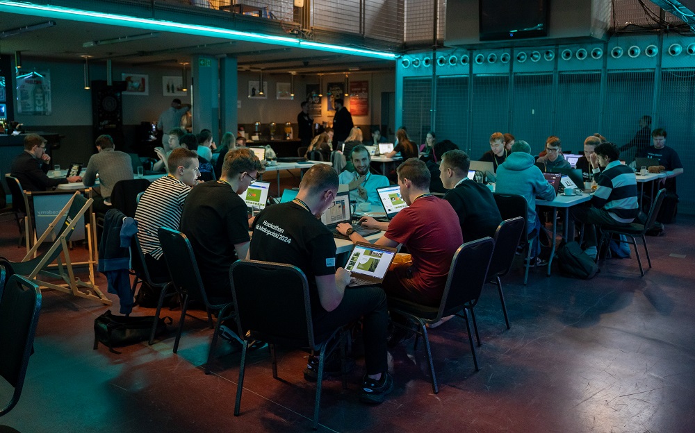zdjęcie z hackathonu przedstawiające młode osoby siedzące przy stołach, pracujące wspólnie, ale każdy ze swoim laptopem