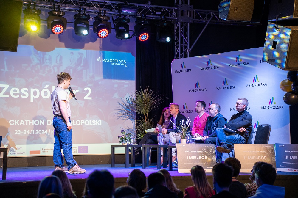 zdjęcie z hackathonu przedstaijace mówcę na stronie zwracjącego się jury