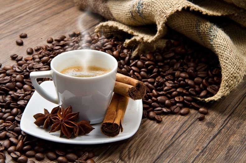 dymiąca filiżanka kawy ustawiona spodku, na którym jest również wanilia i cynamon, w tle z rozsypane z worka ziarna kawy