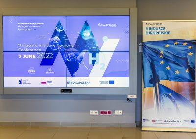 ekran z banerem promującym konferencję, obok rollup informującym o funduszach europejskich w Małopolsce